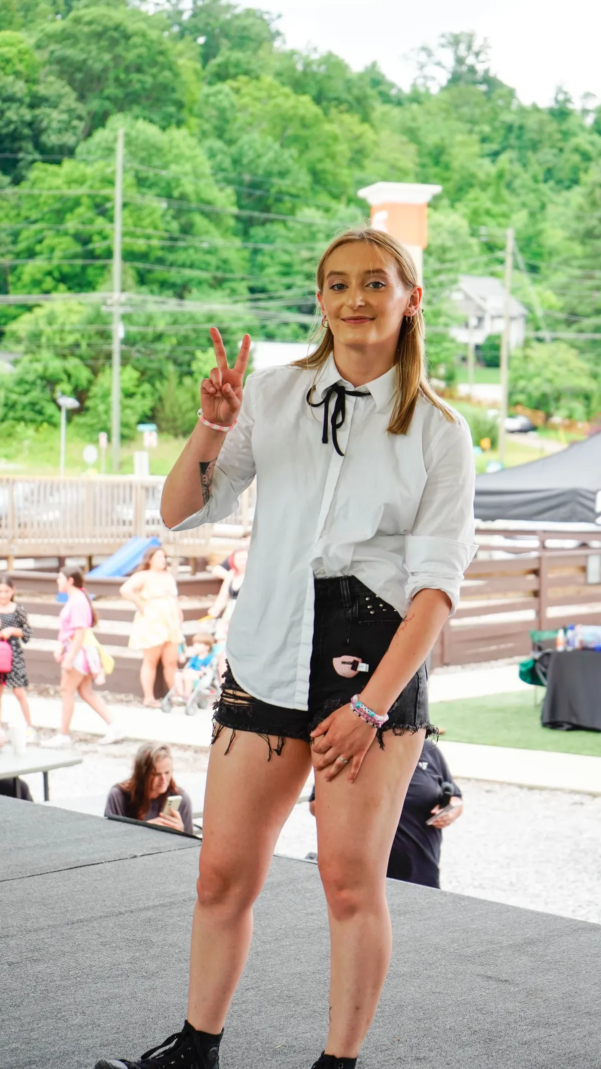 A young woman in a white button-up shirt with a black bow tie and black shorts stands on a stage flashing a peace sign, with a playground and picnic area behind her at Knox Pop Con.